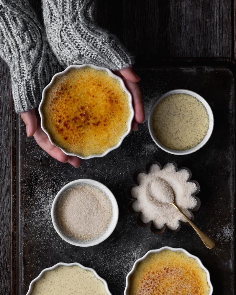 creme brulee on tray with sugar and hands holding one ramekin