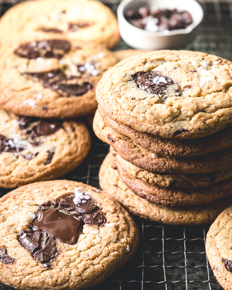 stack of cookies on baking rack surrounded by more cookies with small dish of chocolate and salt in background