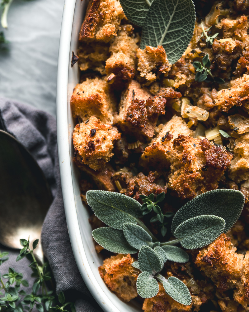 close up of sourdough stuffing with sausage in a white baking dish topped with fresh sage and thyme