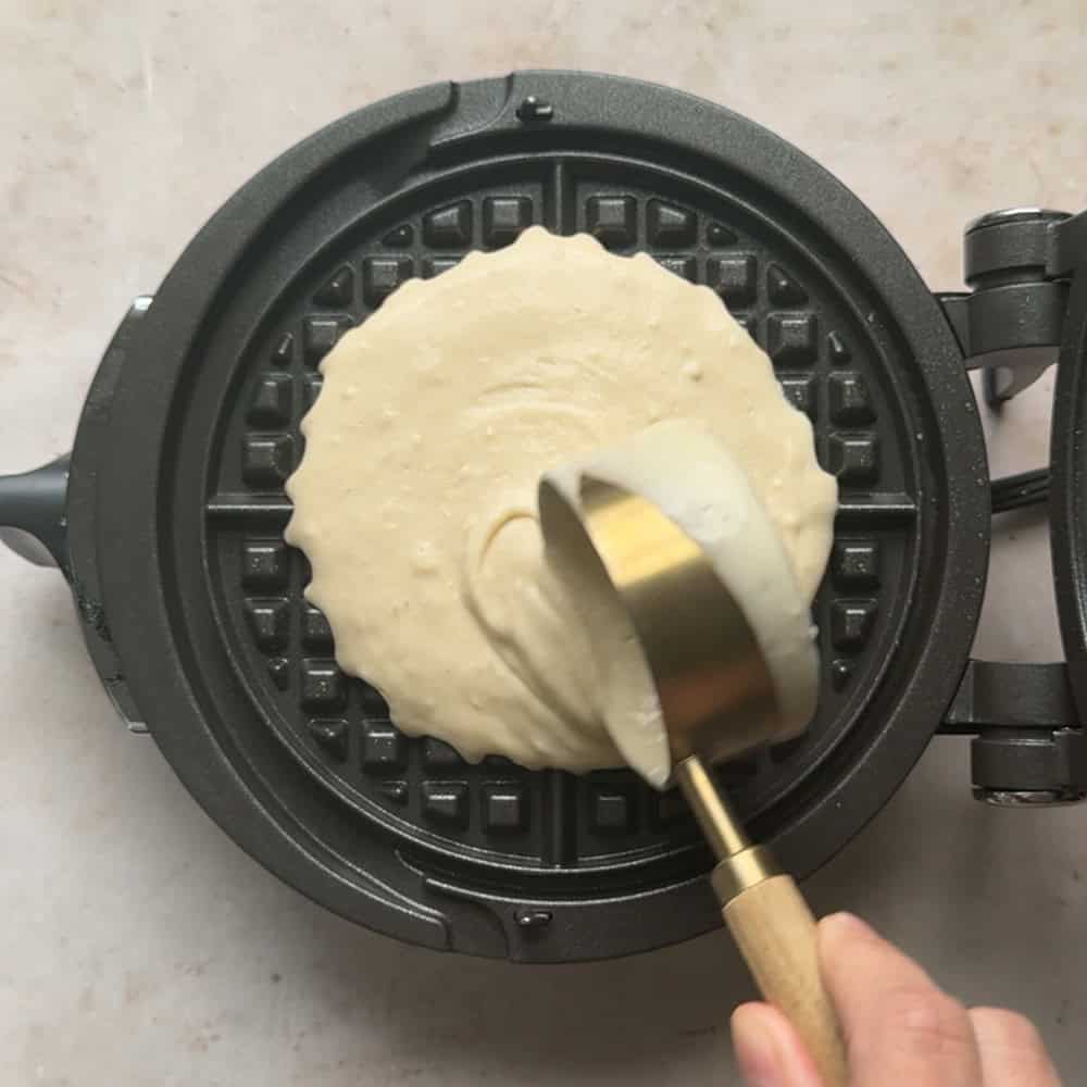 waffle batter being added to waffle iron with a gold measuring cup