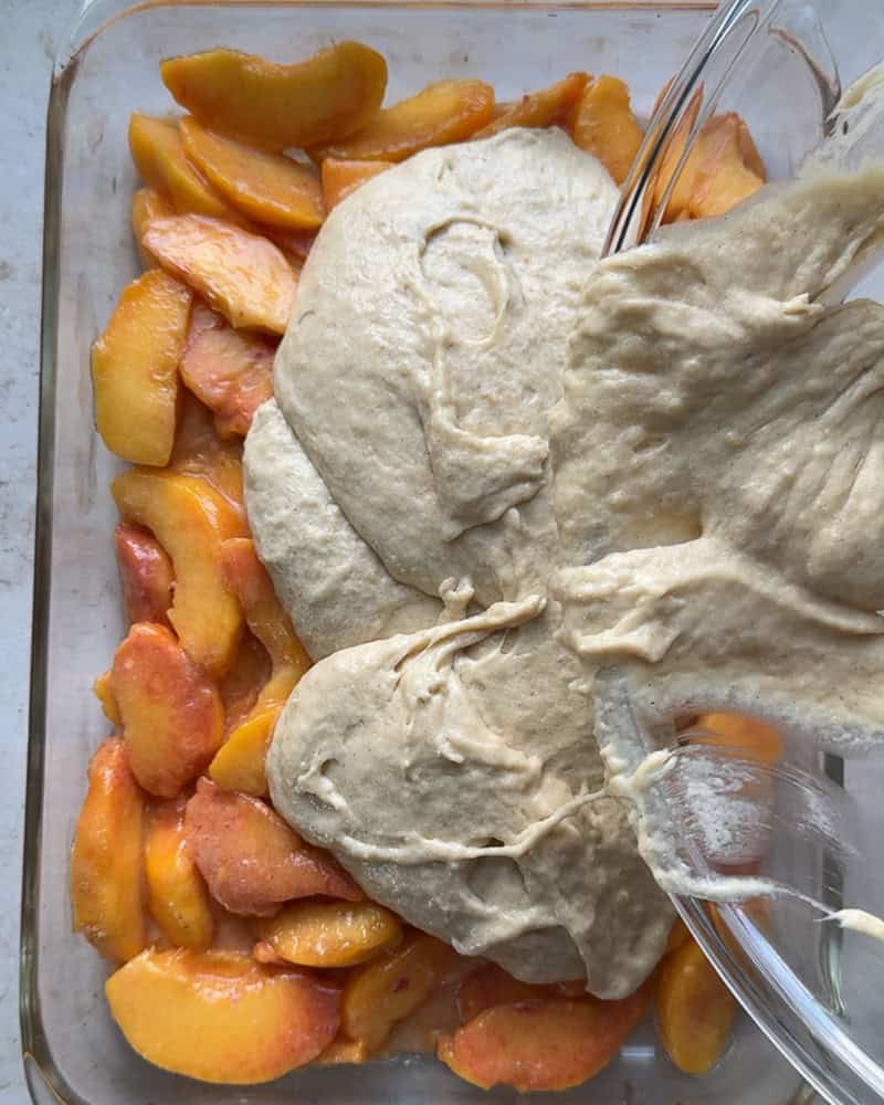 peach cobbler batter being poured onto peach filling in glass baking dish