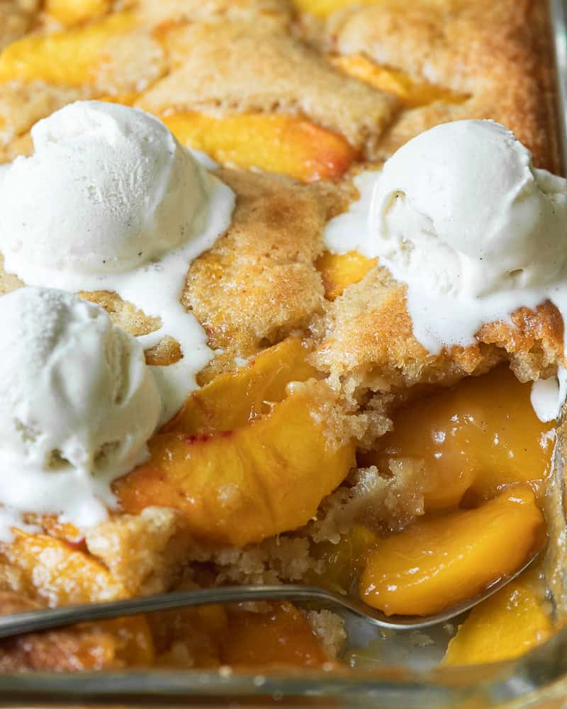 a pan of peach cobbler topped with vanilla ice cream with a spoon holding a peach slice.