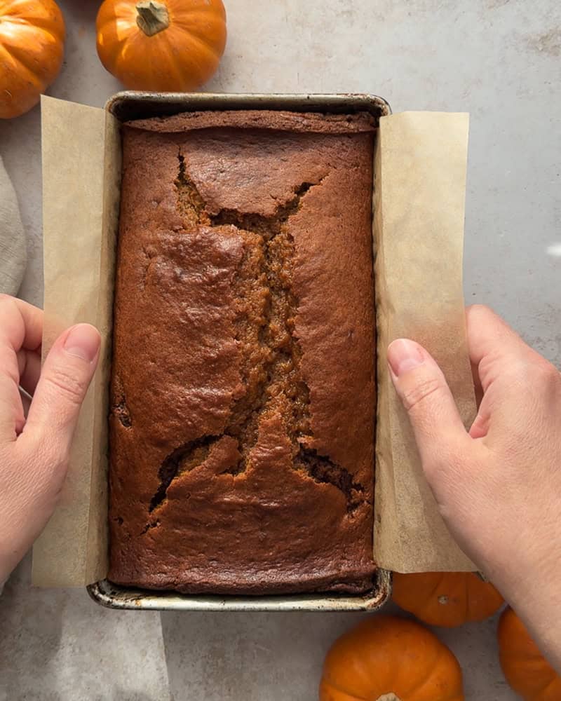 fully baked pumpkin bread being lifted out of pan with parchment paper sling