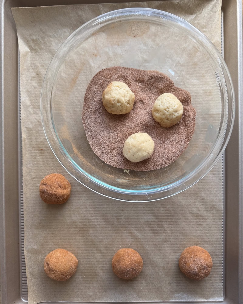 cookie dough balls being rolled in cinnamon sugar and placed onto parchment lined baking sheet