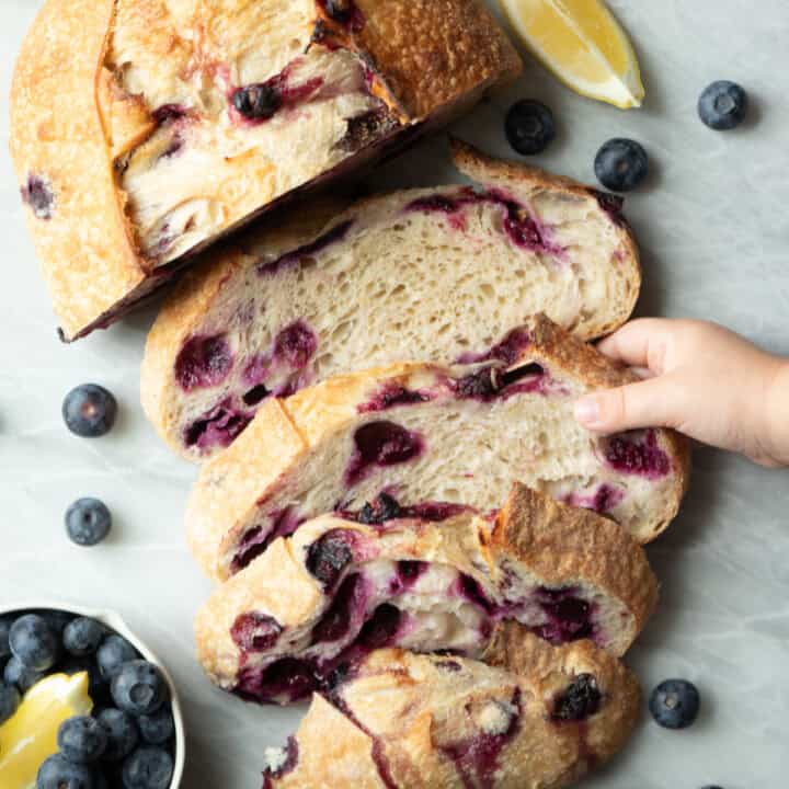 a loaf of sliced blueberry bread with a small hand reaching in grabbing one slice