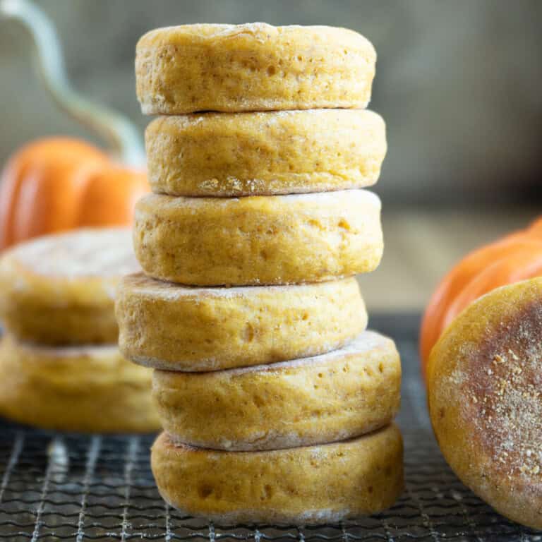 a tall stack of pumpkin English muffins on a wire rack with small pumpkins in the background and foreground