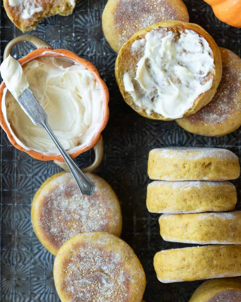 pumpkin english muffins on a wire rack with a small dish of butter. One muffin is split and toasted and topped with butter.