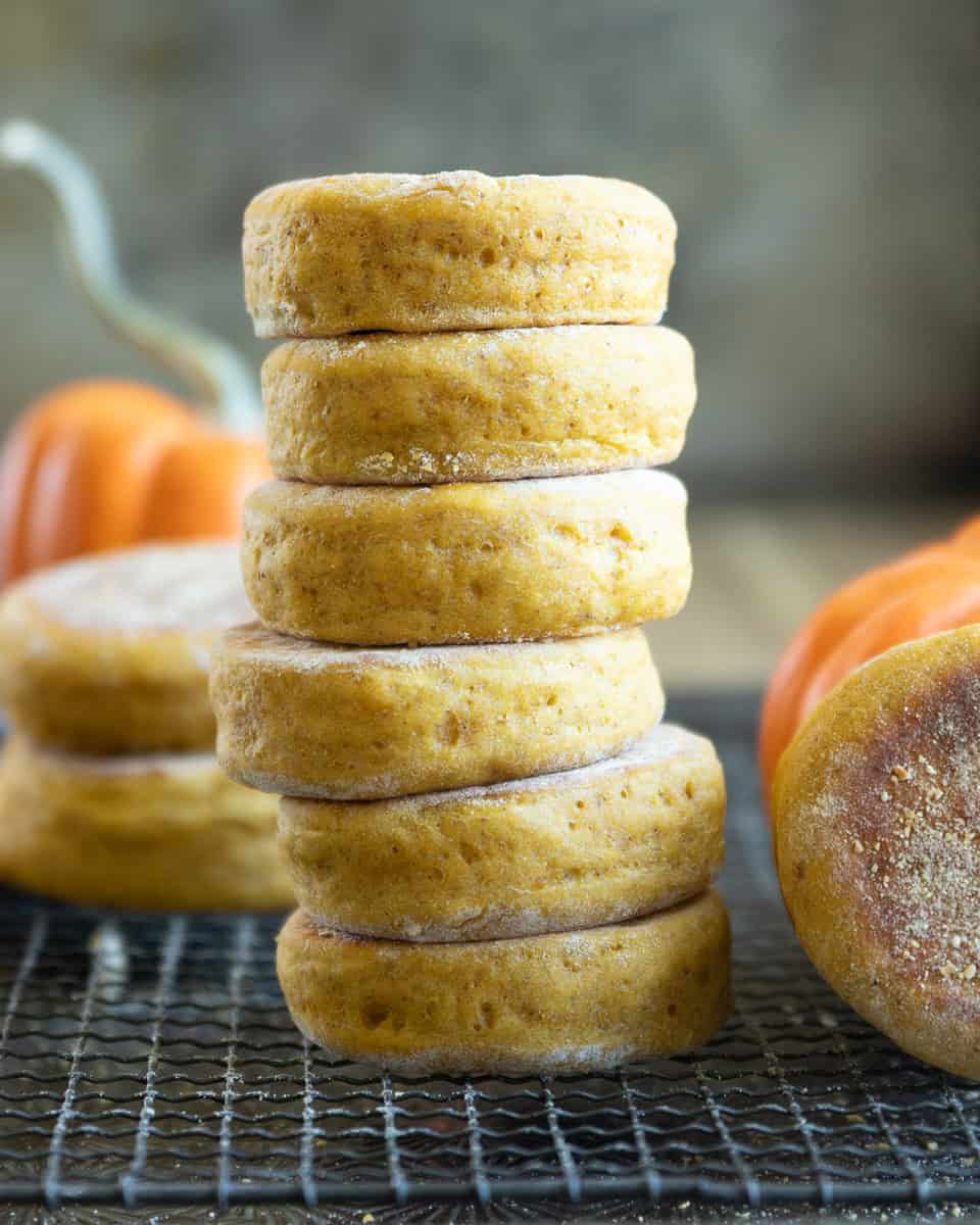 a tall stack of pumpkin English muffins on a wire rack with small pumpkins in the background and foreground